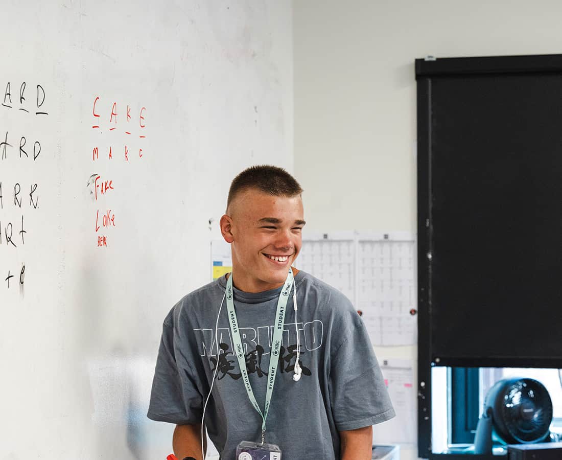 Male student smiling at the front of a Summer Boarding Courses classroom while writing on the board and talking to his friends