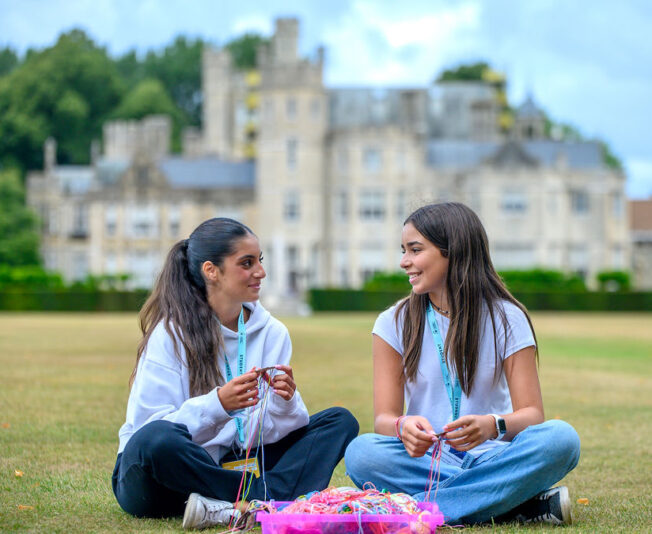 Knitted friendship bracelets, summer camp, outdoor activity, teenage friends, Castle background, summer holiday, bonding activity, relaxed youth, kids crafting, summer learning, outdoor youth groups, creative summer, friendship bands, campus scenery, summer camp fun.