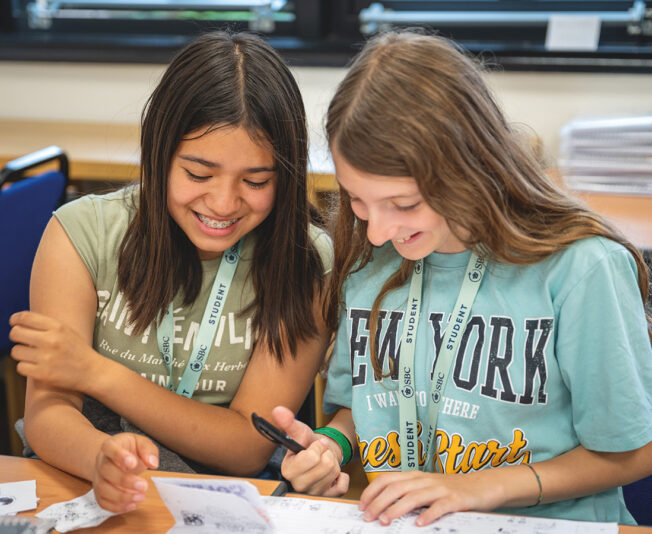 Female students smiling and sat together at a desk in an SBC Canford classroom working on some class work together