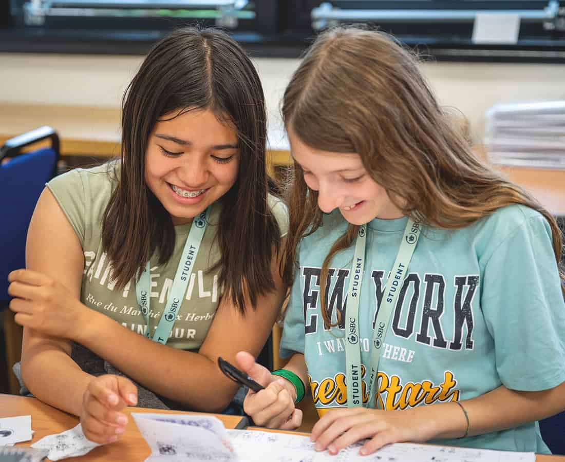 Female students smiling and sat together at a desk in an SBC Canford classroom working on some class work together