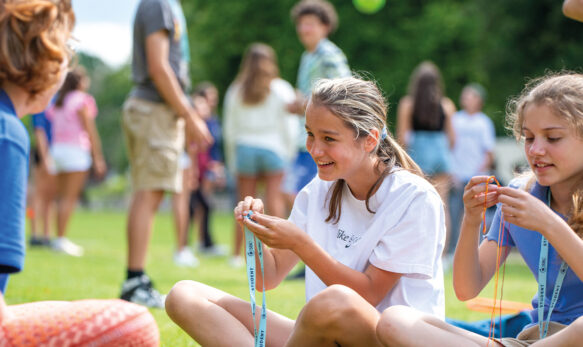 Two female students sat with a member of staff at SBC Canford Summer School, they are outside sat on the grass smiling and chatting