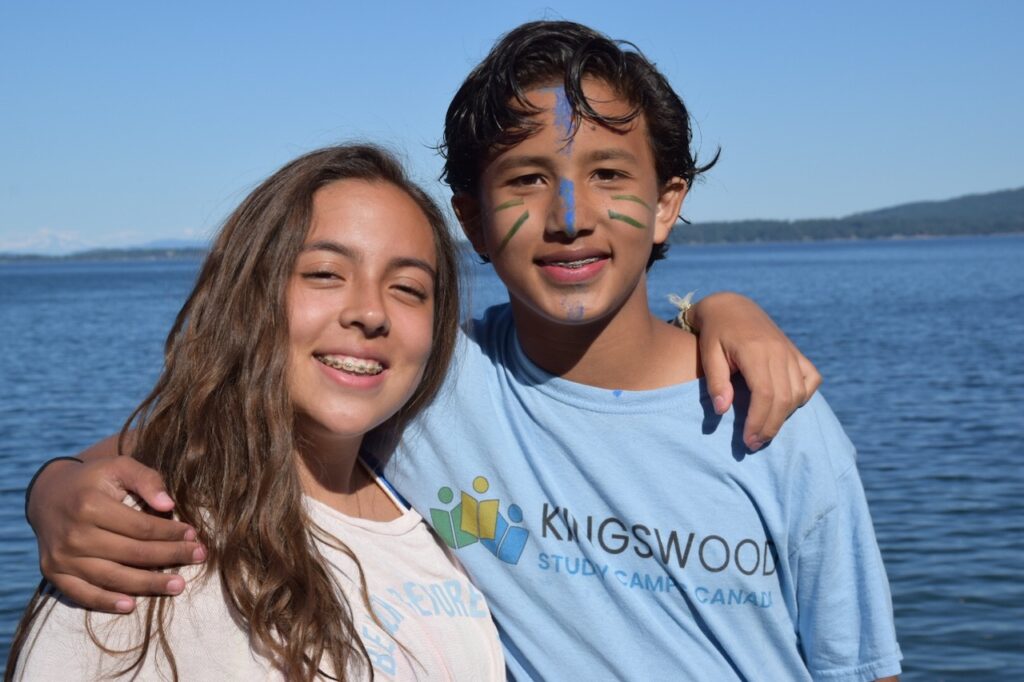 Two campers with face paint stand arm in arm beside a lake in Kingswood t-shirts