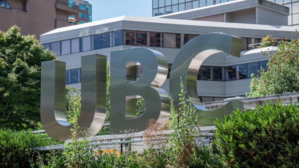 Large UBC letters stand outside a modern glass campus building among green plants