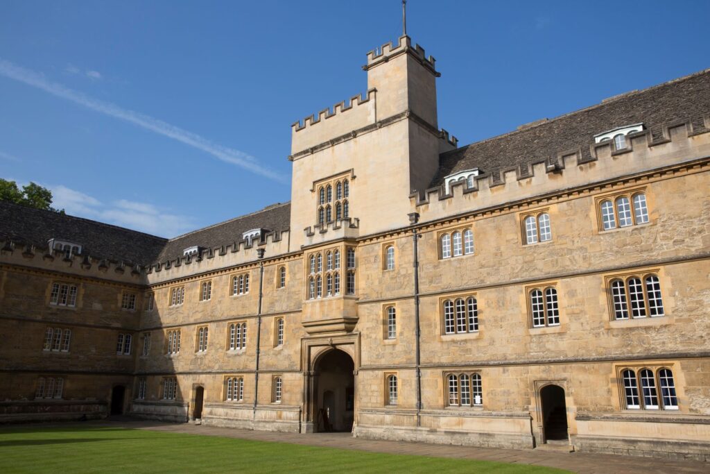 A honey coloured stone college quad with a battlemented tower and green lawn