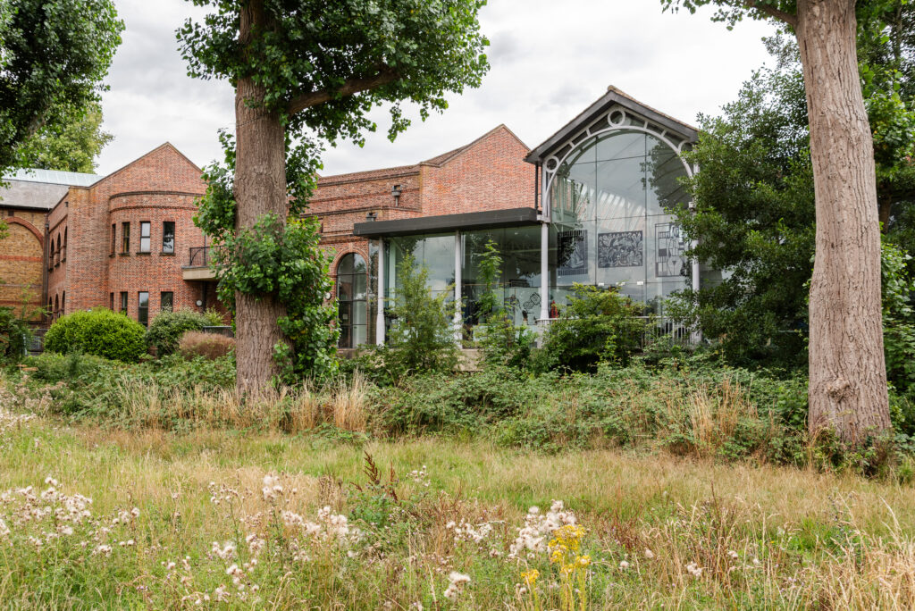 Modern glass-fronted educational building surrounded by greenery and mature trees in a lush outdoor setting.