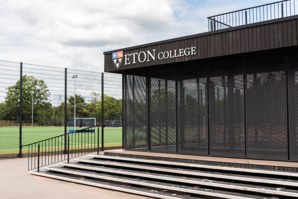 Modern school building with Eton College logo, outdoor sports facilities, rugby field, and trees in the background