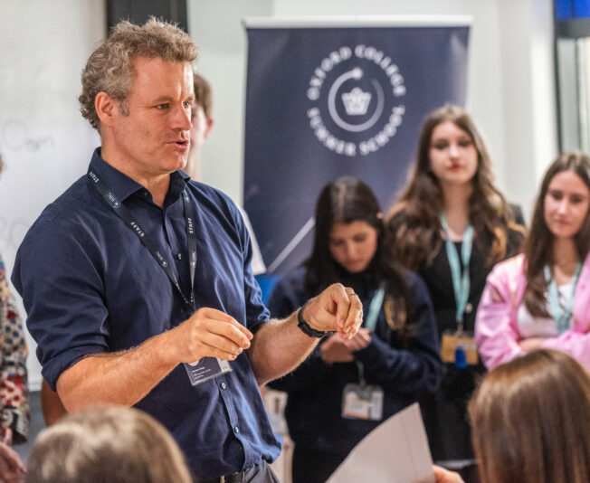 A teacher demonstrates a task to students beside an Oxford College Summer School banner