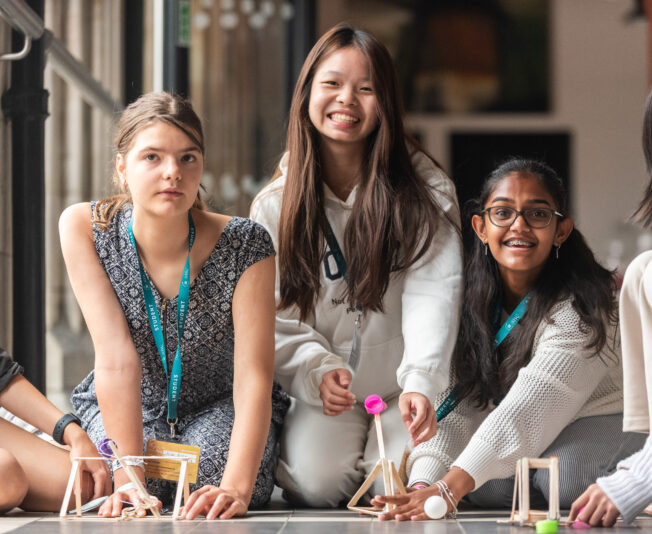 Students sit outdoors building small towers from sticks and marshmallows during a group task