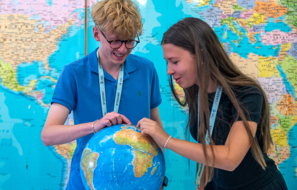 Two students point at a globe in front of a large wall world map